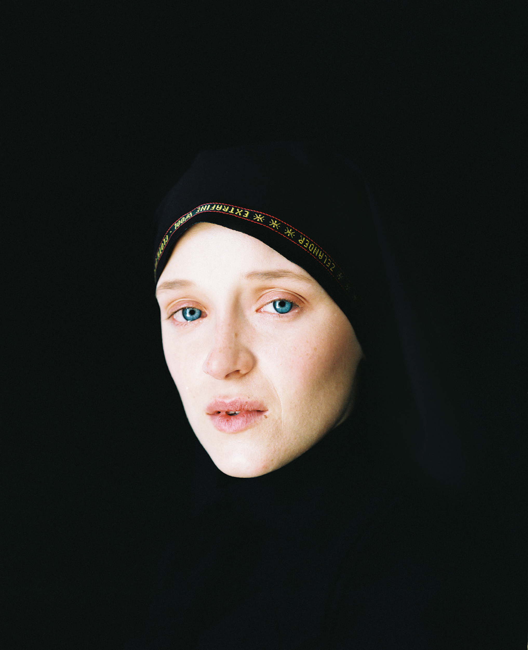 Close-up studio portrait of a woman with blue eyes wearing a black head covering with gold trim, set against a dark black background.