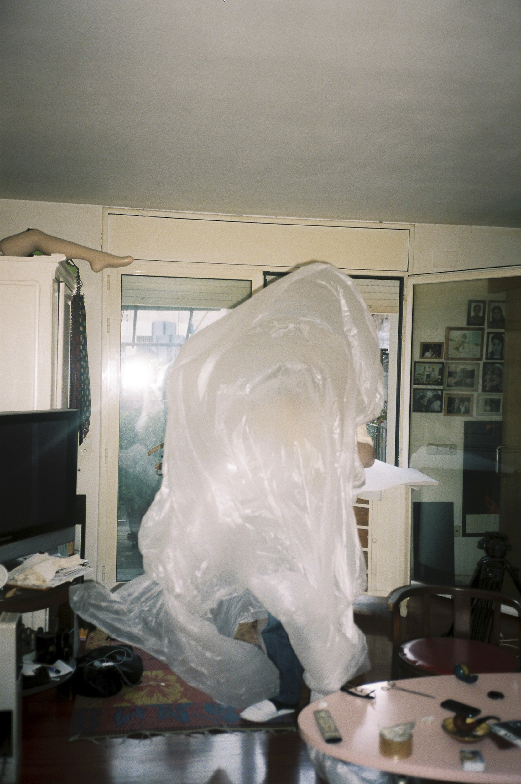 Person in a translucent plastic sheet stands indoors among home furnishings, illuminated by a doorway in a candid-style photo.