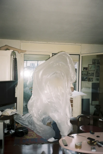 Person in a translucent plastic sheet stands indoors among home furnishings, illuminated by a doorway in a candid-style photo.
