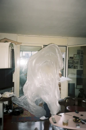 Person in a translucent plastic sheet stands indoors among home furnishings, illuminated by a doorway in a candid-style photo.