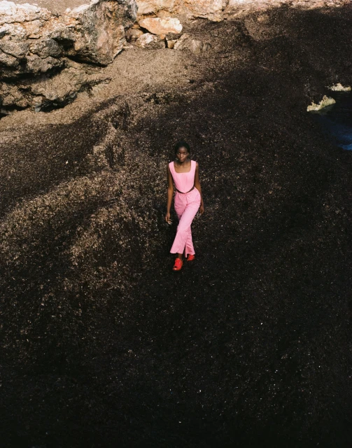 A woman in a pink outfit confidently strides across a dark, textured landscape towards a rocky coastline on a sunny day.