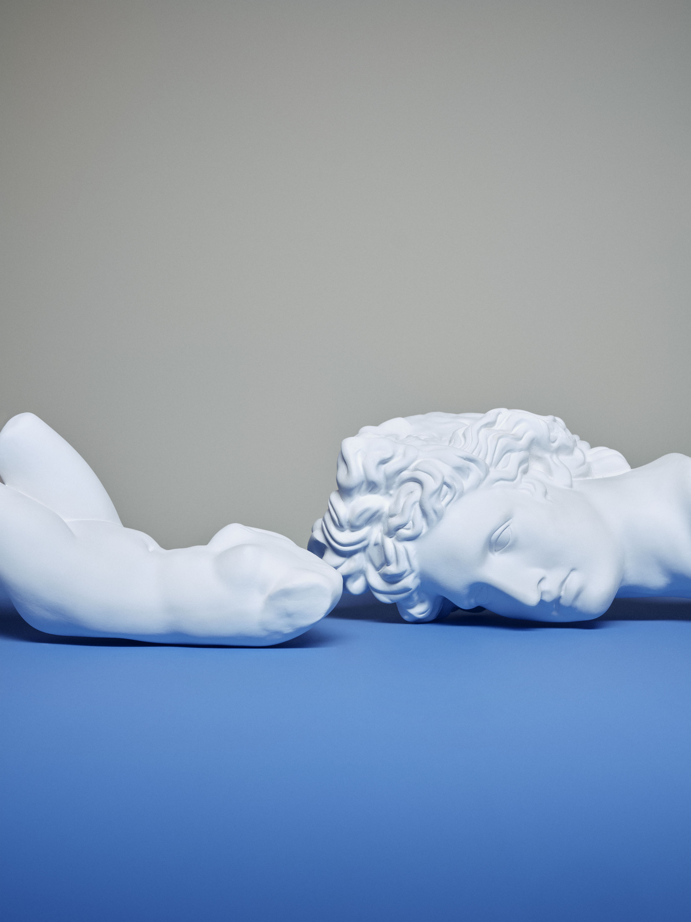 Two white plaster sculptures of human bodies on a blue surface with a grey background. Classical figures, studio lighting, minimalist aesthetic.