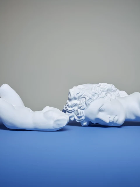 Two white plaster sculptures of human bodies on a blue surface with a grey background. Classical figures, studio lighting, minimalist aesthetic.