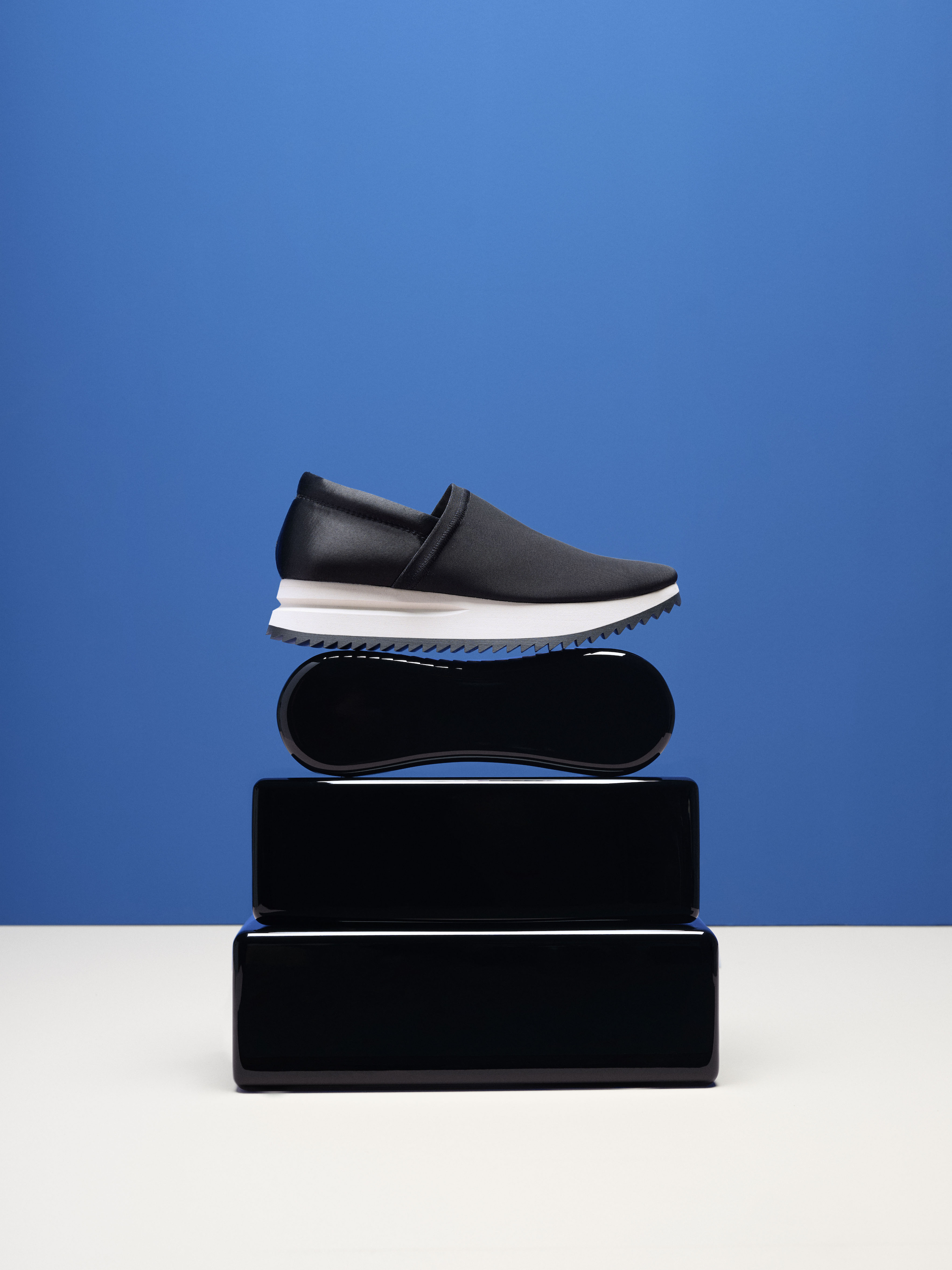 Studio shot of a black slip-on shoe with a white sole stacked on black rectangular risers against a royal blue backdrop.