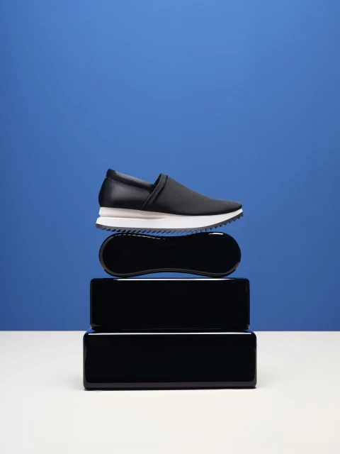 Studio shot of a black slip-on shoe with a white sole stacked on black rectangular risers against a royal blue backdrop.