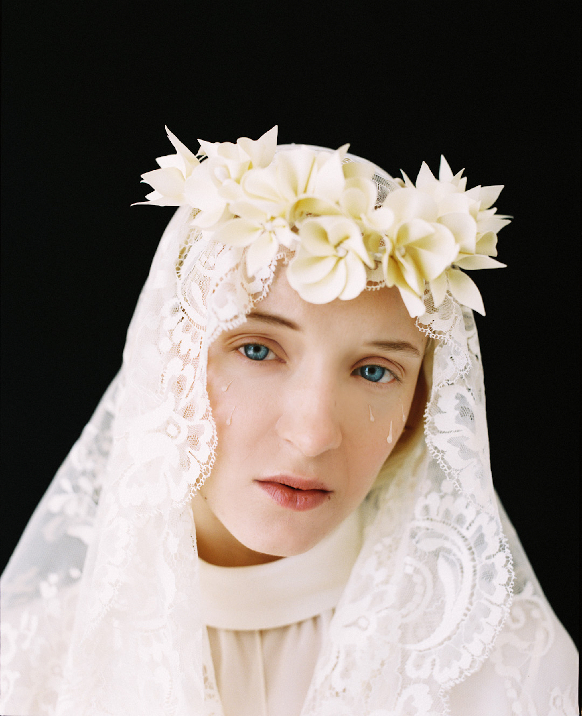 Close-up of a woman with fair skin and blue eyes wearing a white floral crown and lace veil, tear-like droplets on her cheek.