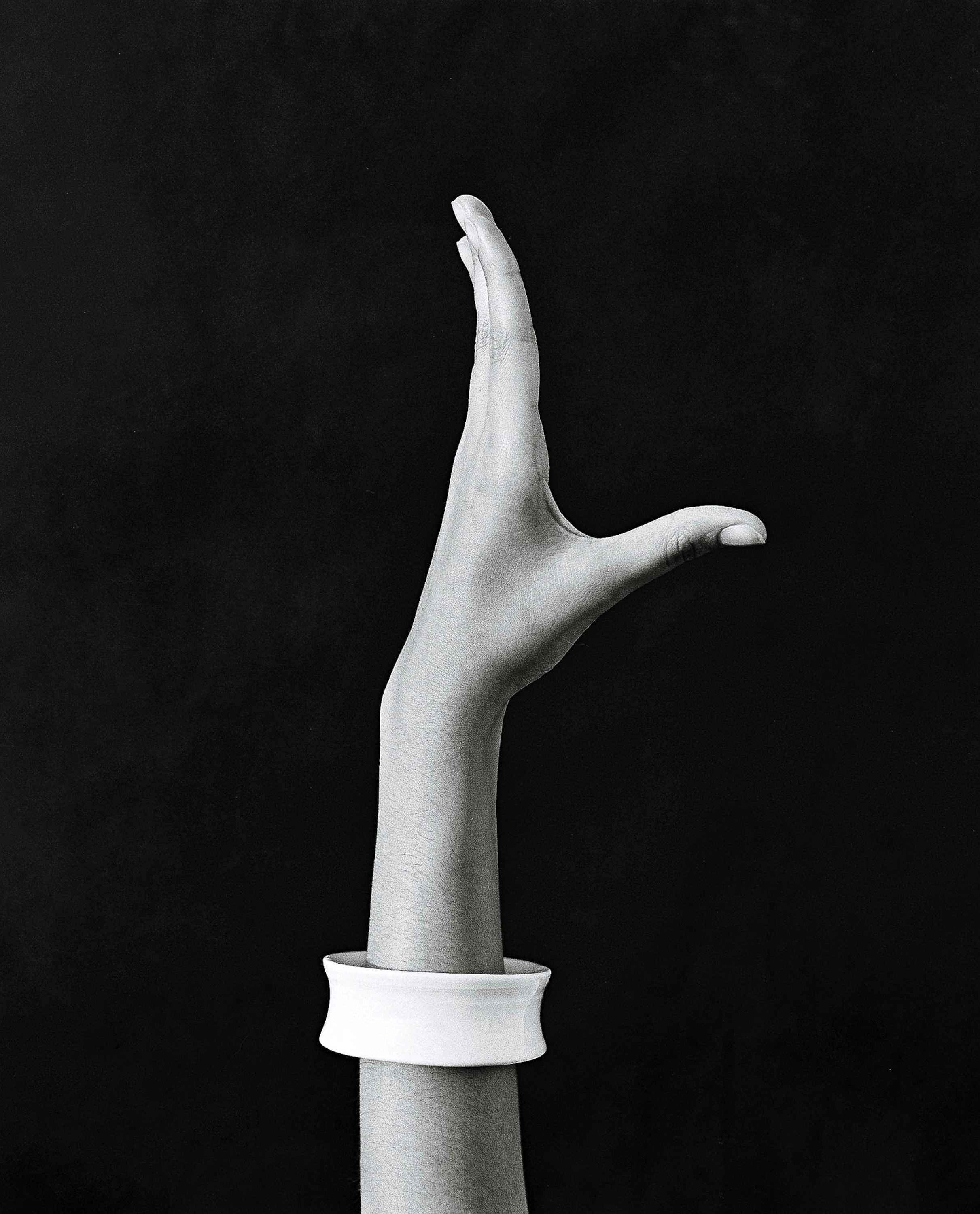 Monochrome portrait of a raised hand wearing a white cuff bracelet against a dark background by Rita Puig-serra Costa