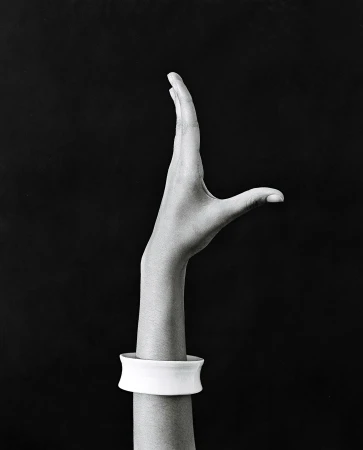 Monochrome portrait of a raised hand wearing a white cuff bracelet against a dark background by Rita Puig-serra Costa
