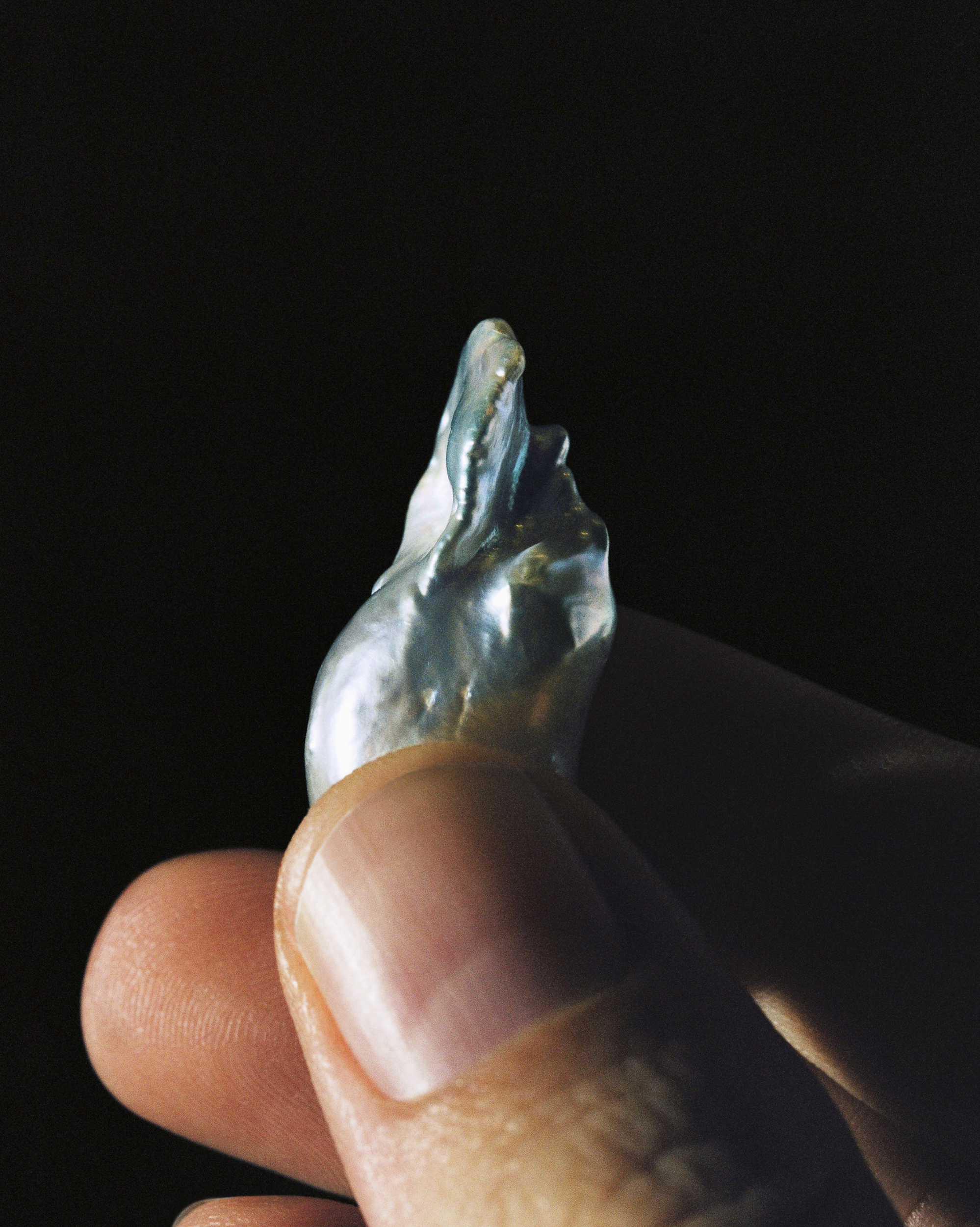 Hand holds baroque pearl with iridescent shimmer against a dark background, close-up studio shot for jewelry or fashion.