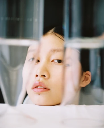 Close-up studio shot of an Asian young adult woman looking at the camera through clear glass panels creating distortions.