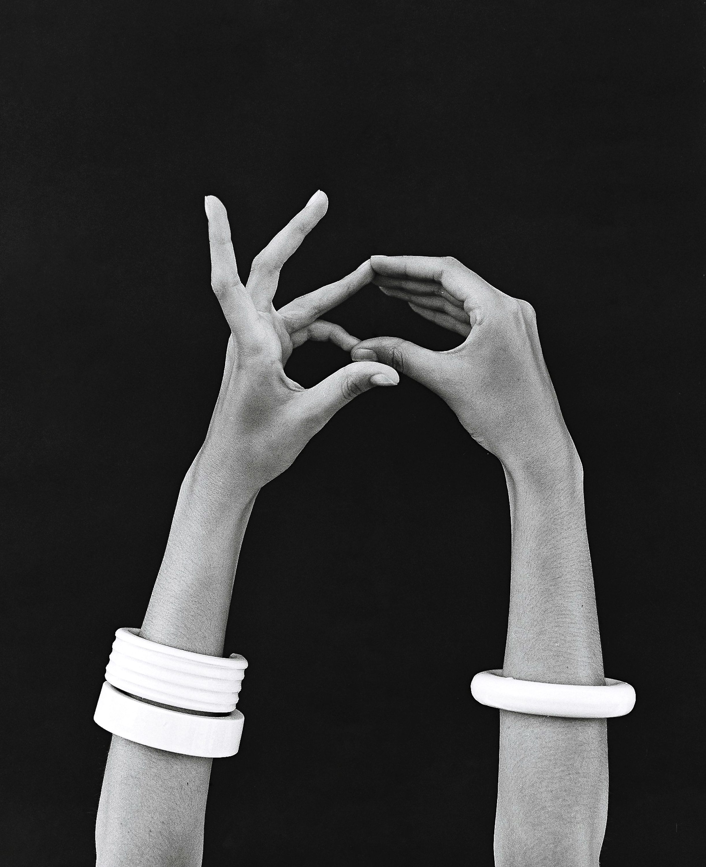 Monochromatic studio shot of dark-skinned hands forming a triangle, adorned with white bangles, against a black background.