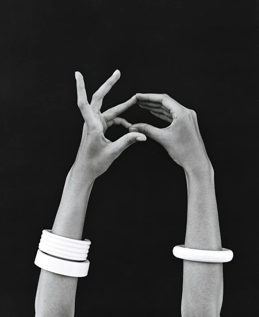 Monochromatic studio shot of dark-skinned hands forming a triangle, adorned with white bangles, against a black background.