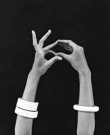 Monochromatic studio shot of dark-skinned hands forming a triangle, adorned with white bangles, against a black background.