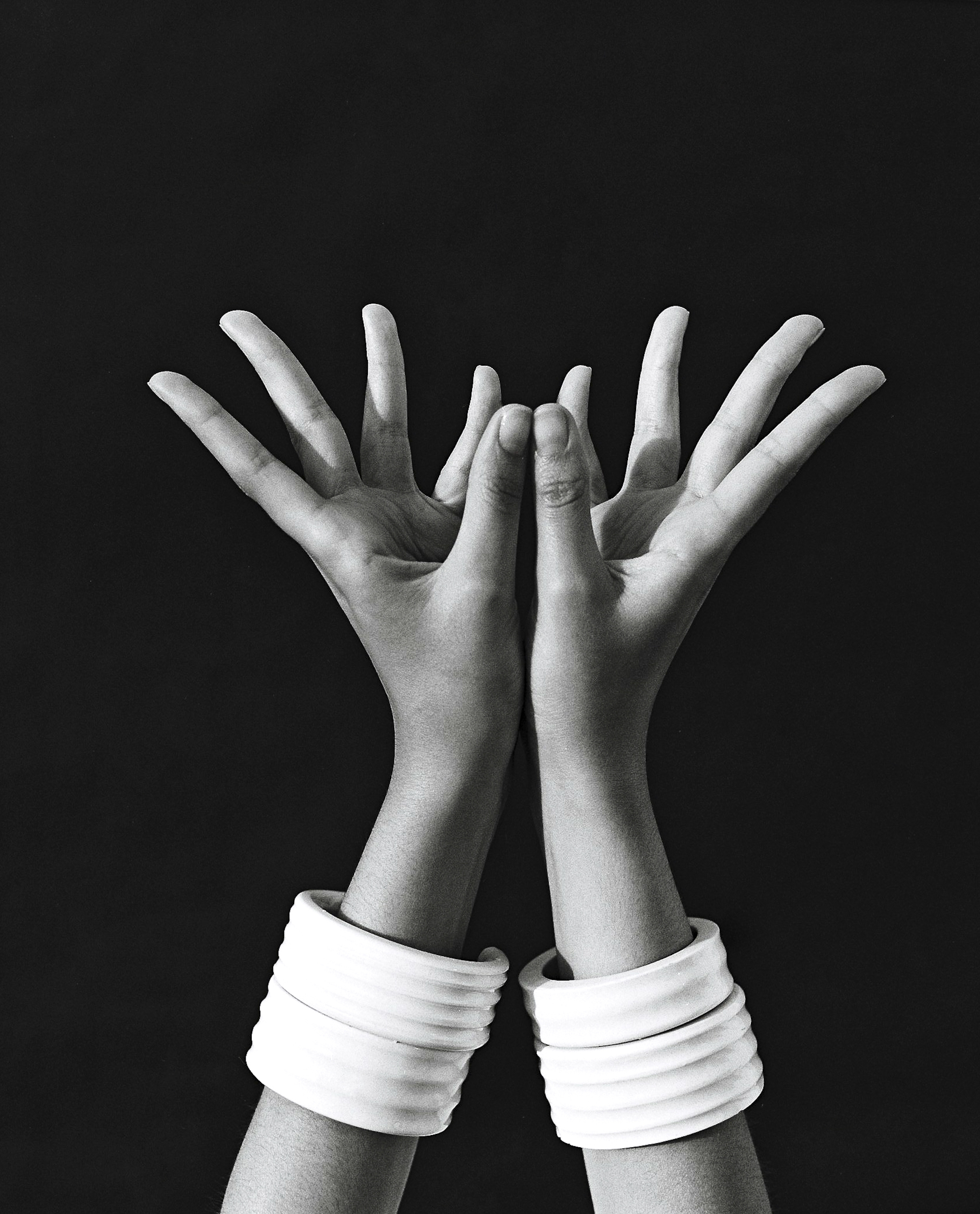Monochrome studio shot of two arms raised with hands open, adorned with white, ribbed bracelets against a stark black background.