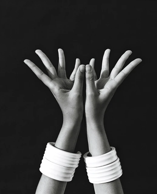 Monochrome studio shot of two arms raised with hands open, adorned with white, ribbed bracelets against a stark black background.