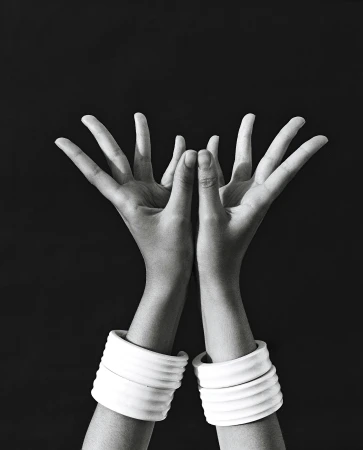 Monochrome studio shot of two arms raised with hands open, adorned with white, ribbed bracelets against a stark black background.