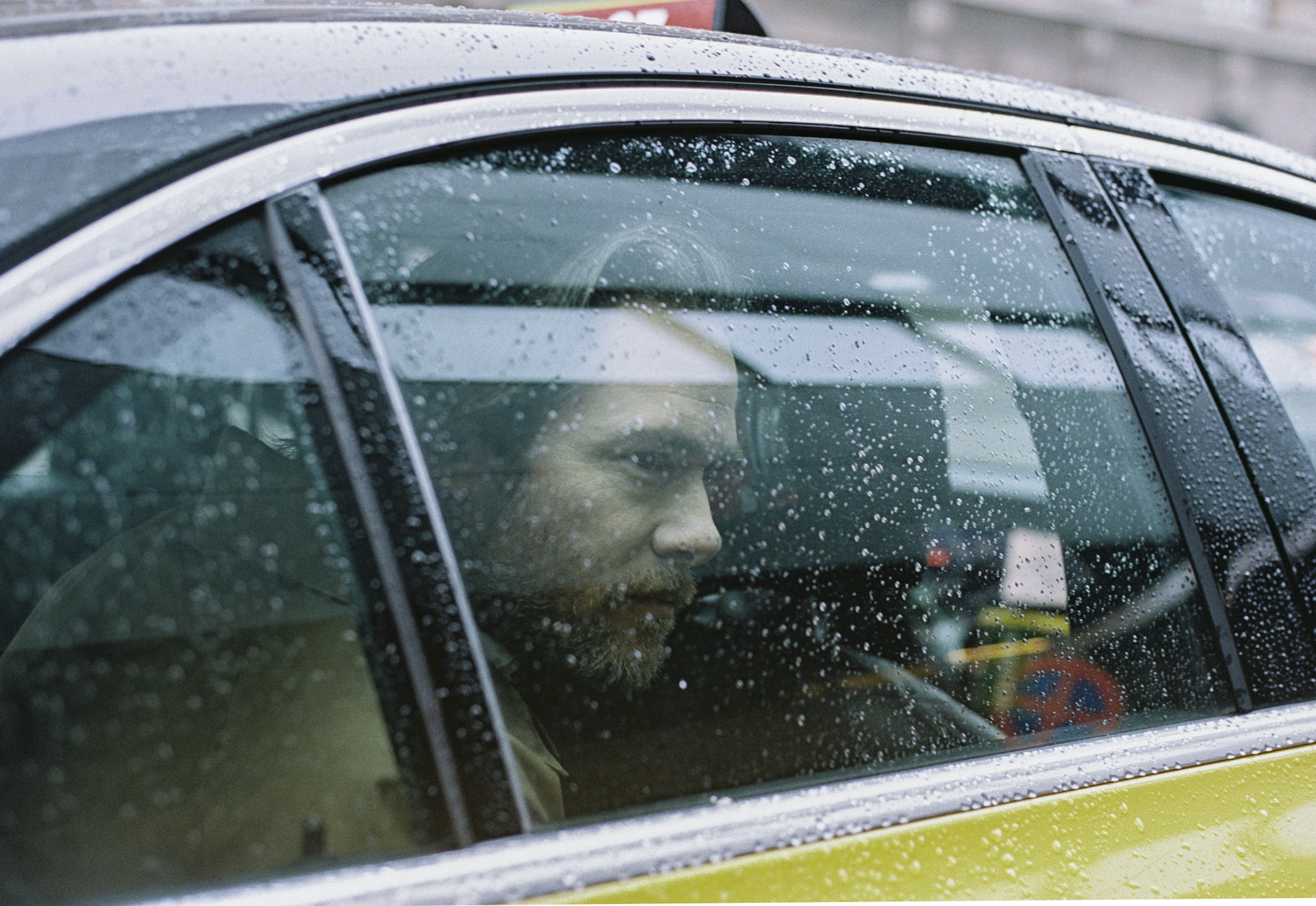 Adult man with beard sits in the back of a yellow taxi on a rainy day, viewed through the water-streaked window.