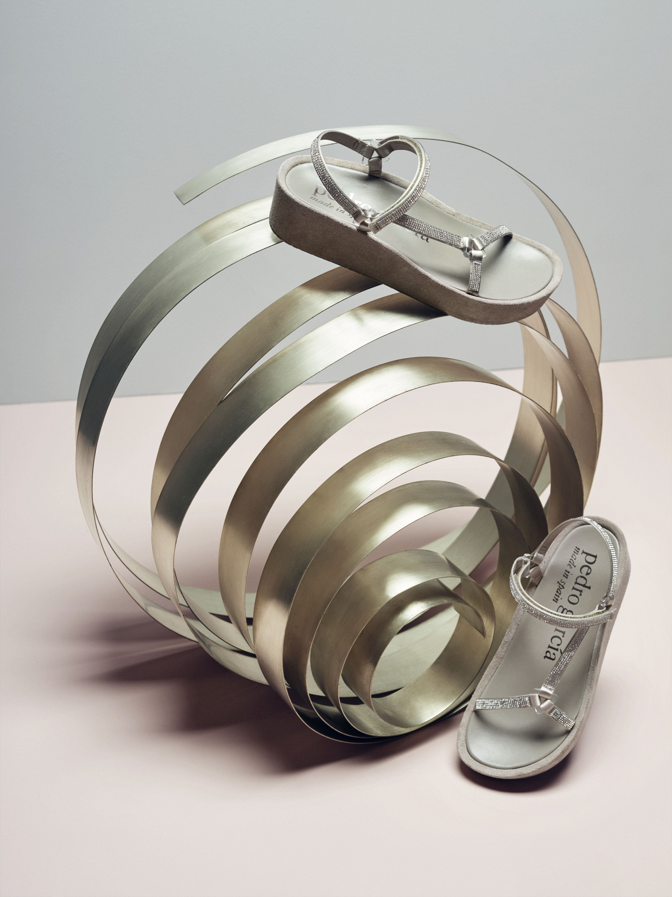 Elevated shot of platform sandals arranged alongside a metal sculpture, set against a two-tone grey and light pink background.