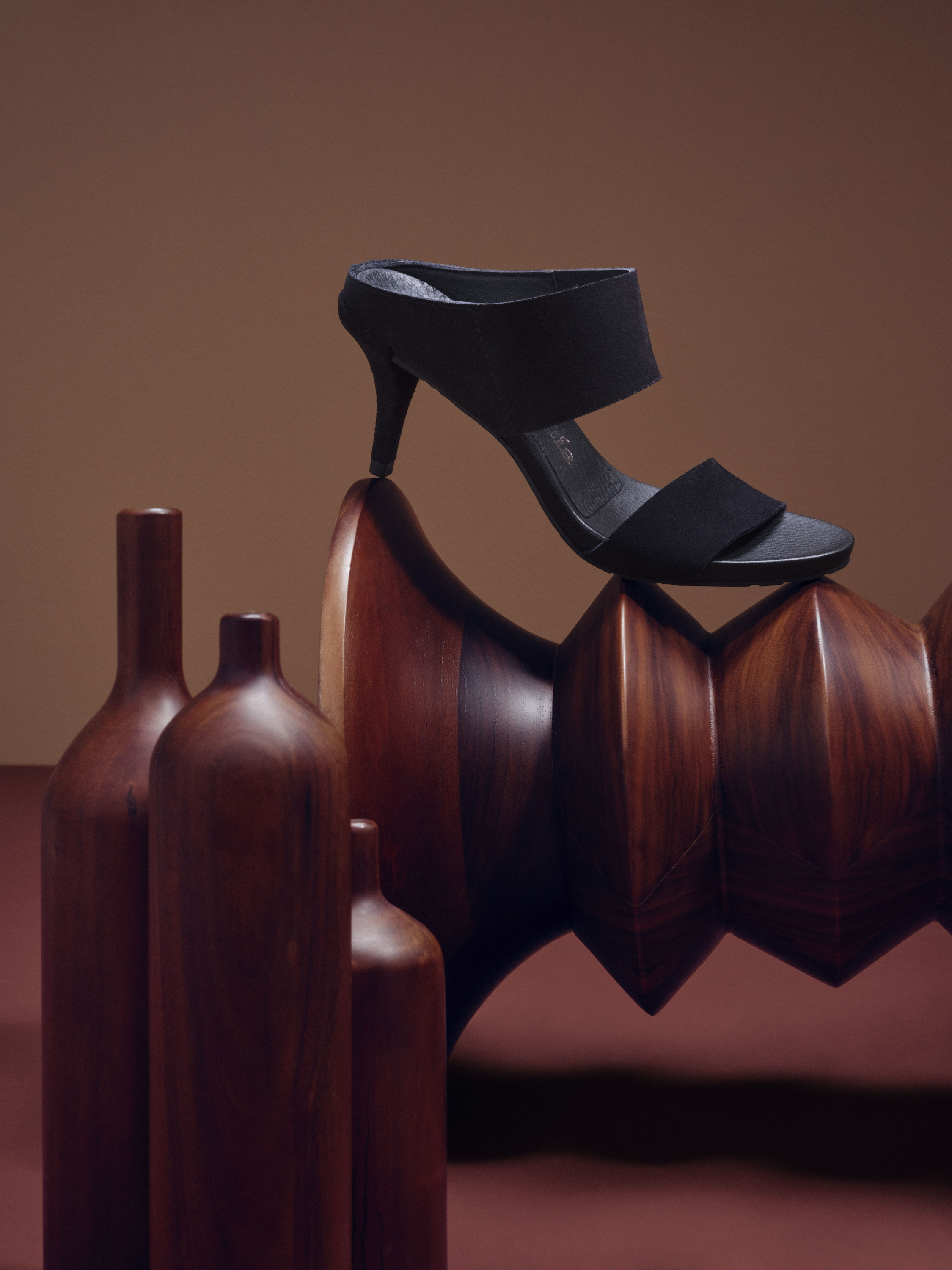 Black heeled sandal on a wooden sculpture next to dark brown bottles, set against a warm brown background.