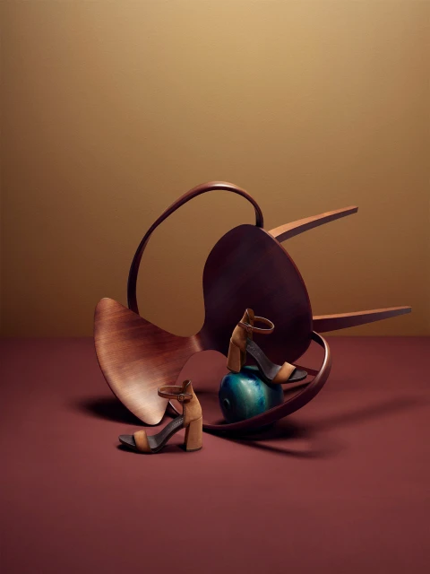 Still life of tan block-heeled sandals on a green sphere with a wooden chair on a maroon surface against a tan background.