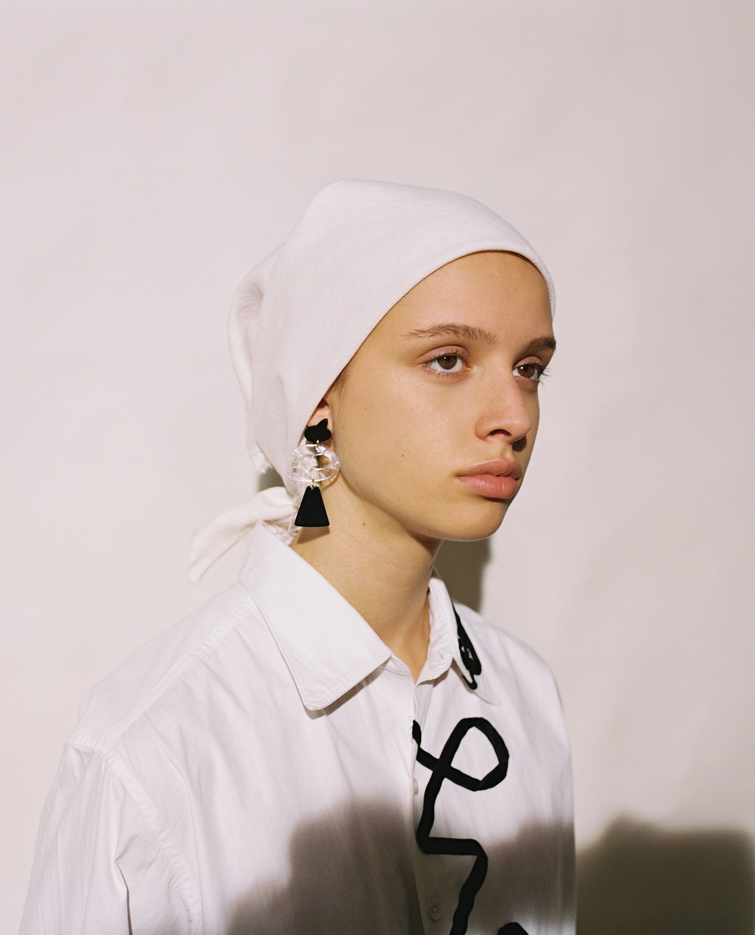Portrait of a young woman wearing a white headscarf, statement earrings, and a decorated white shirt against a white background.