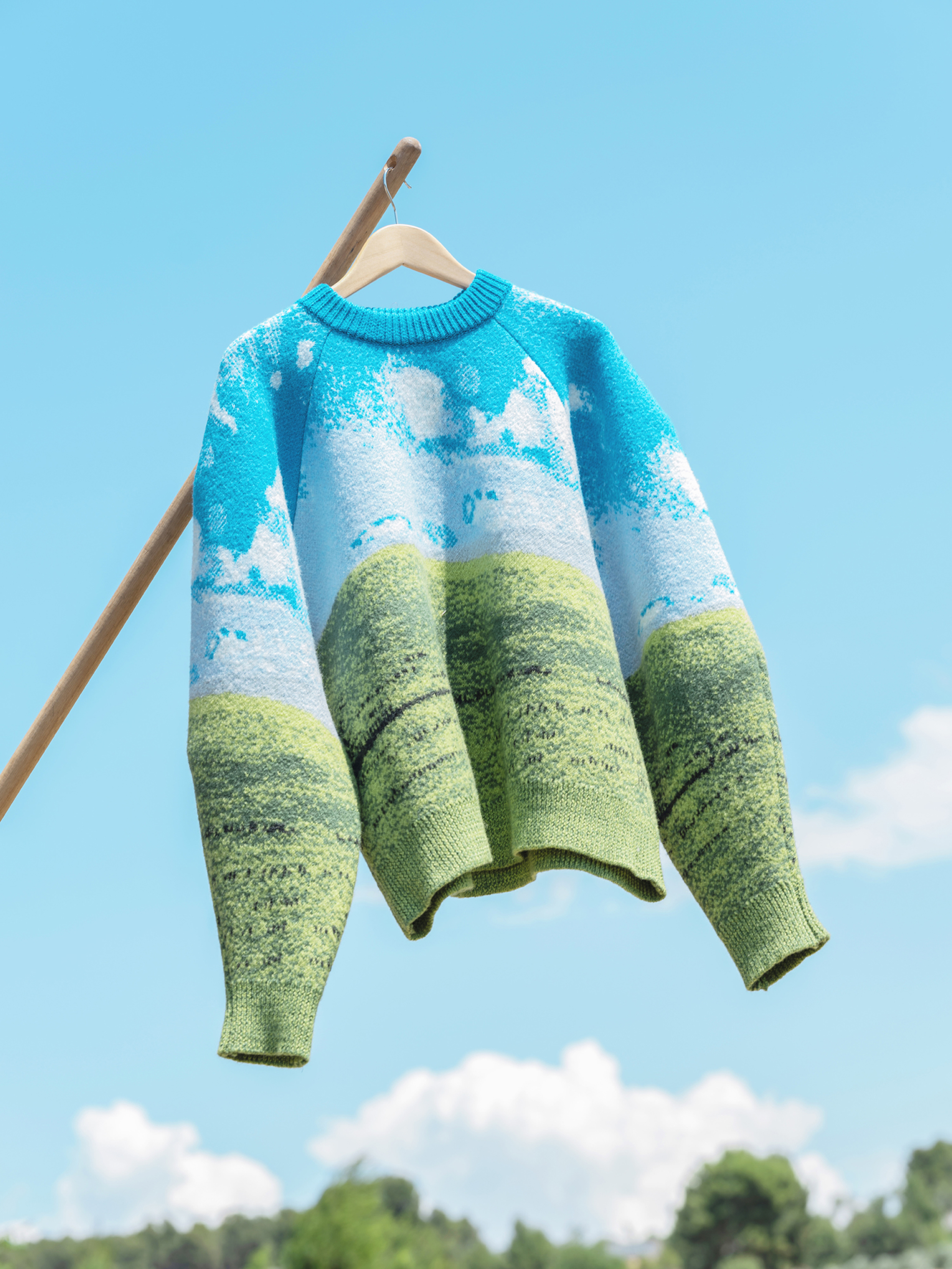 Sweater with landscape design on a hanger, set against a bright blue sky and distant treeline, creating a whimsical outdoor apparel shot.
