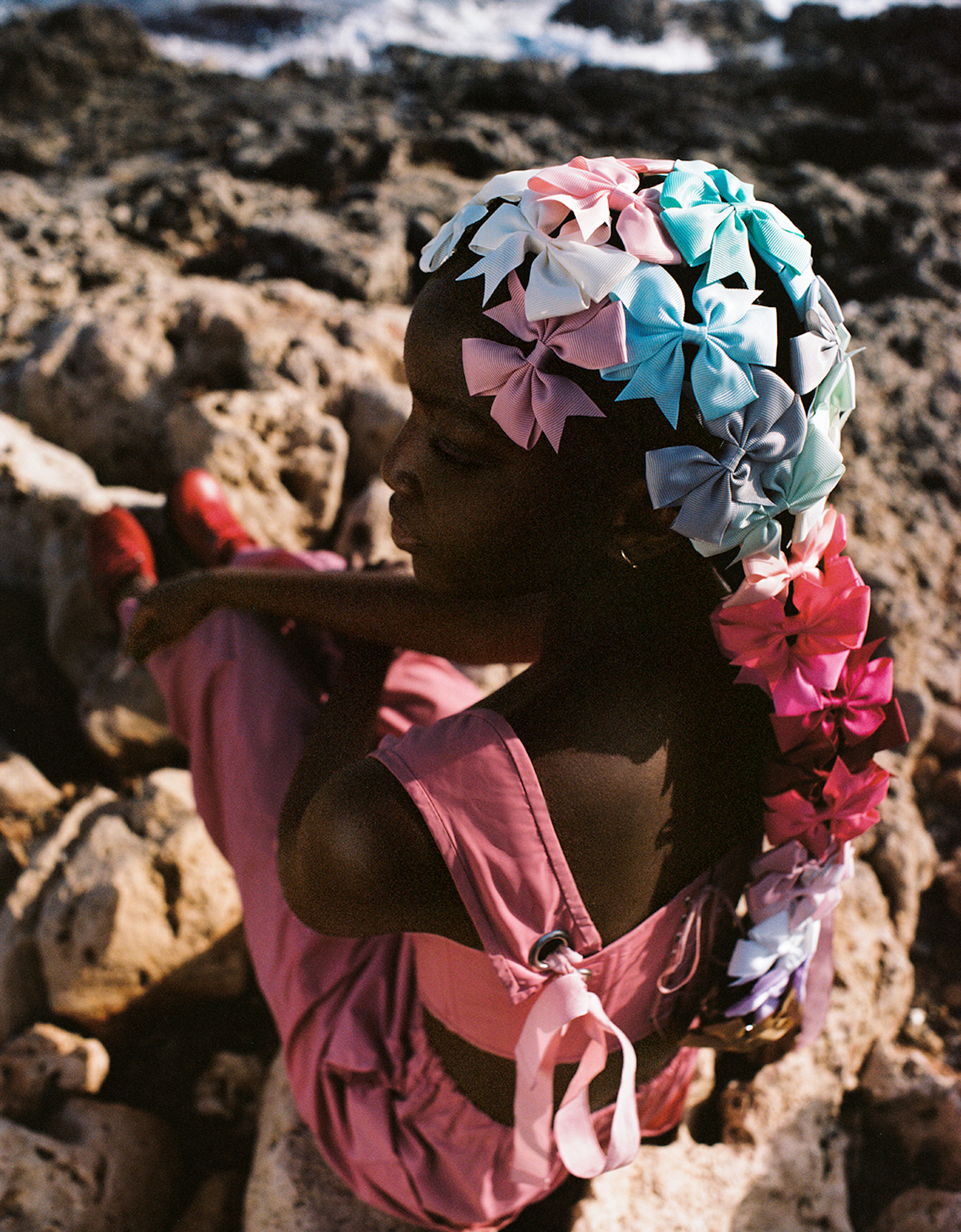 Young girl poses on rocky beach wearing a pink jumpsuit and a colorful hairpiece made of assorted ribbons and bows in outdoor lighting.