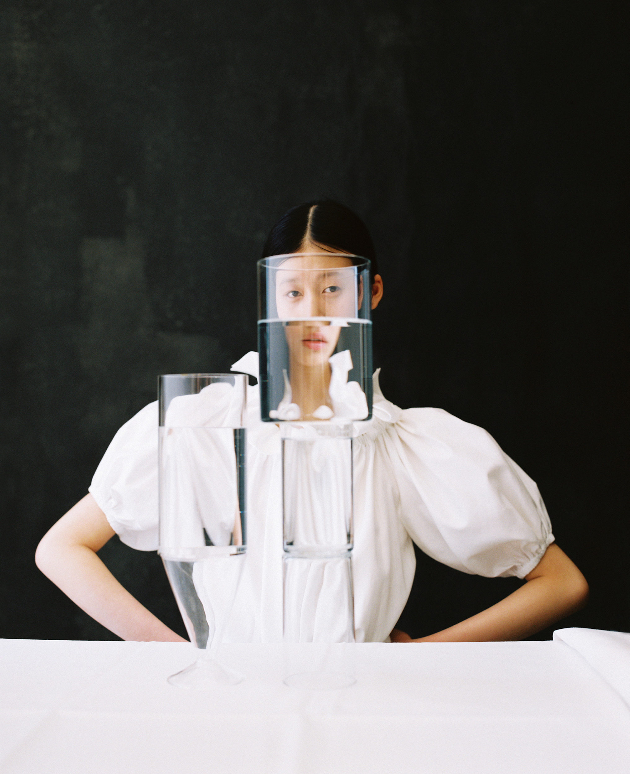 Conceptual fashion shot of an Asian woman behind stacked glasses filled with water, wearing a puffy-sleeved white top against a dark backdrop.