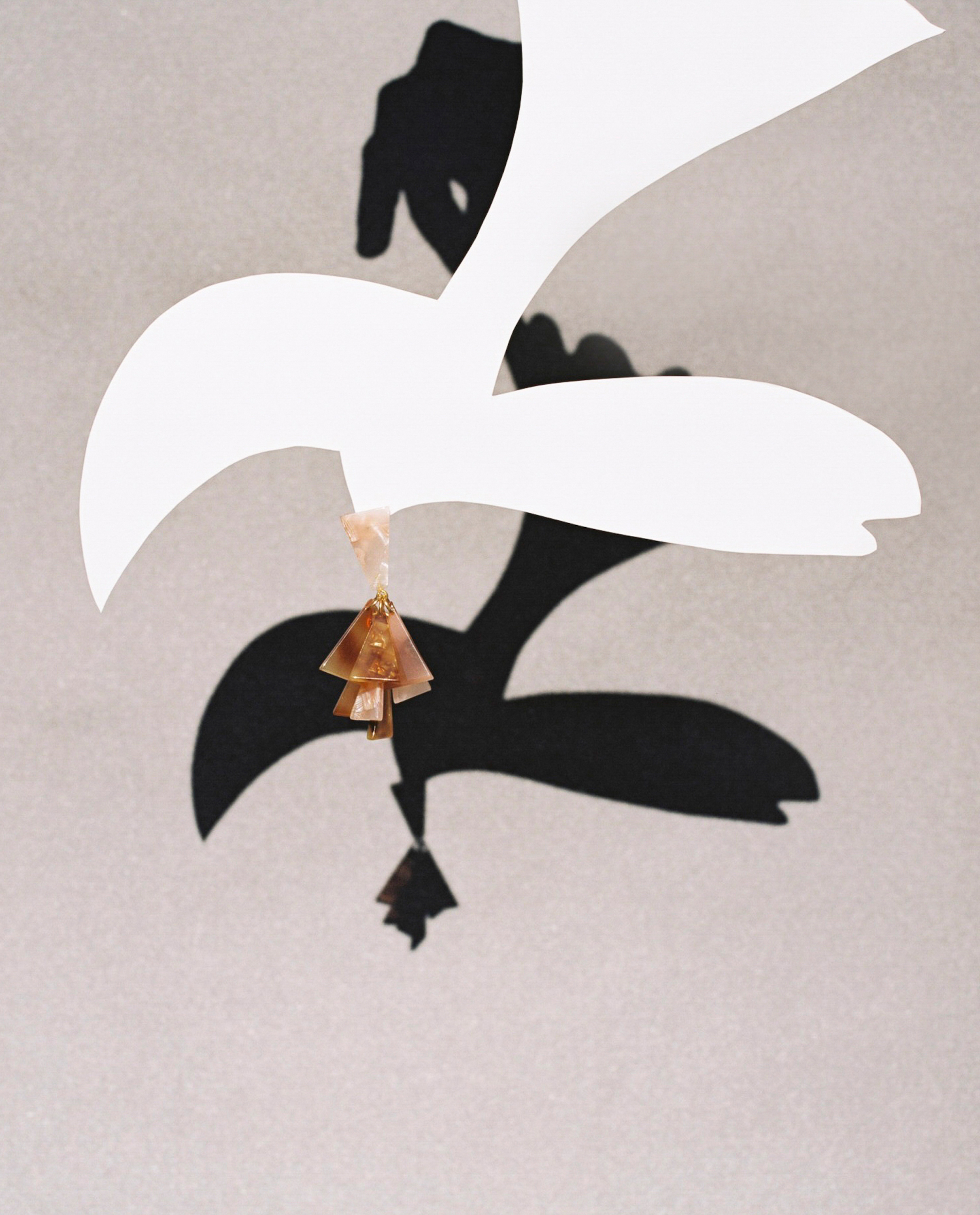 White paper bird cutout casting shadow on textured wall, adorned with brown beaded pendant. Minimalist art with clean aesthetic.