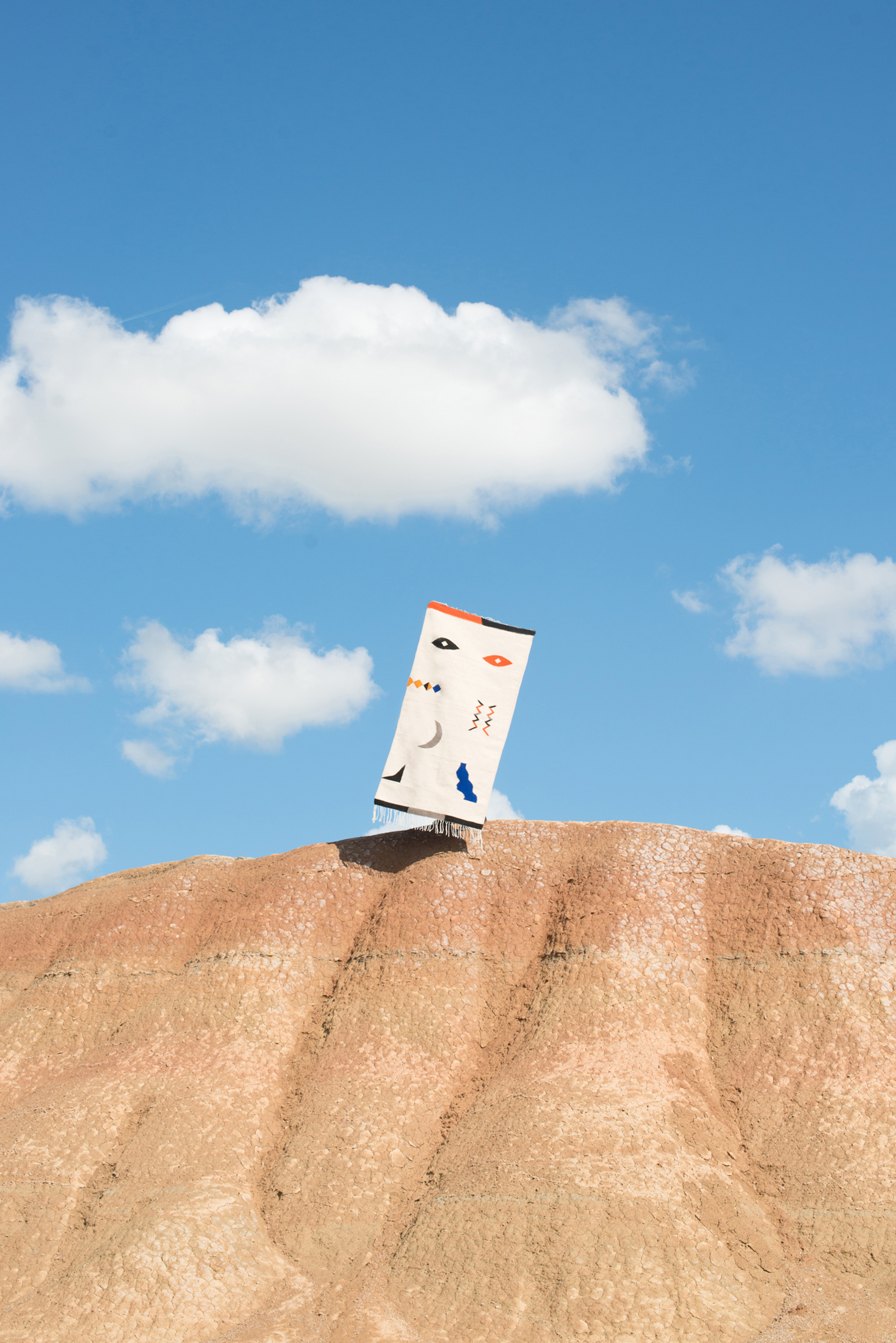 Abstract artwork on a placard placed on a desert hill under a blue sky with clouds. The artwork features symbolic facial elements.