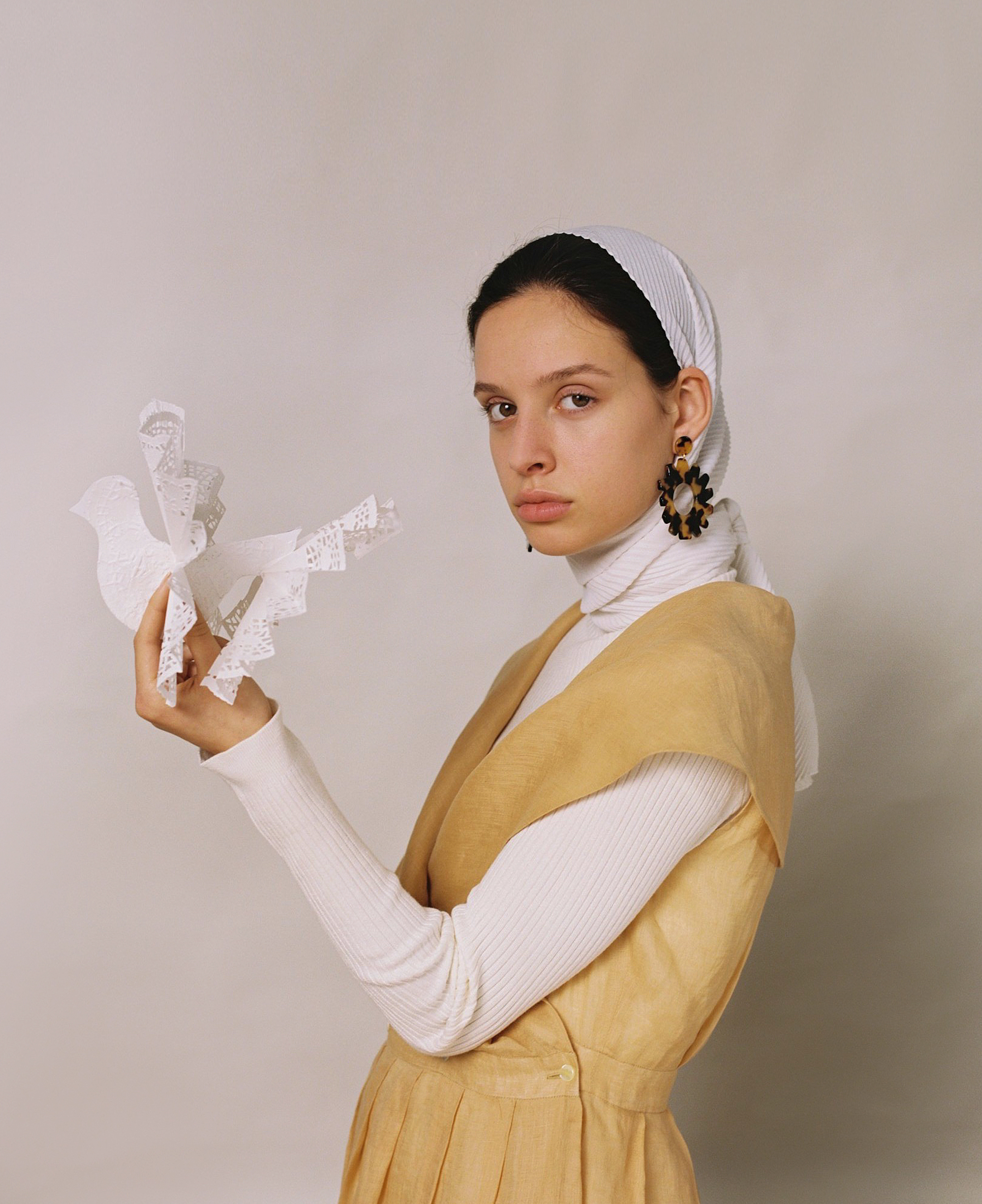 Portrait of a young woman in a light-yellow dress and white headscarf holding a decorative paper bird against a light-grey background.