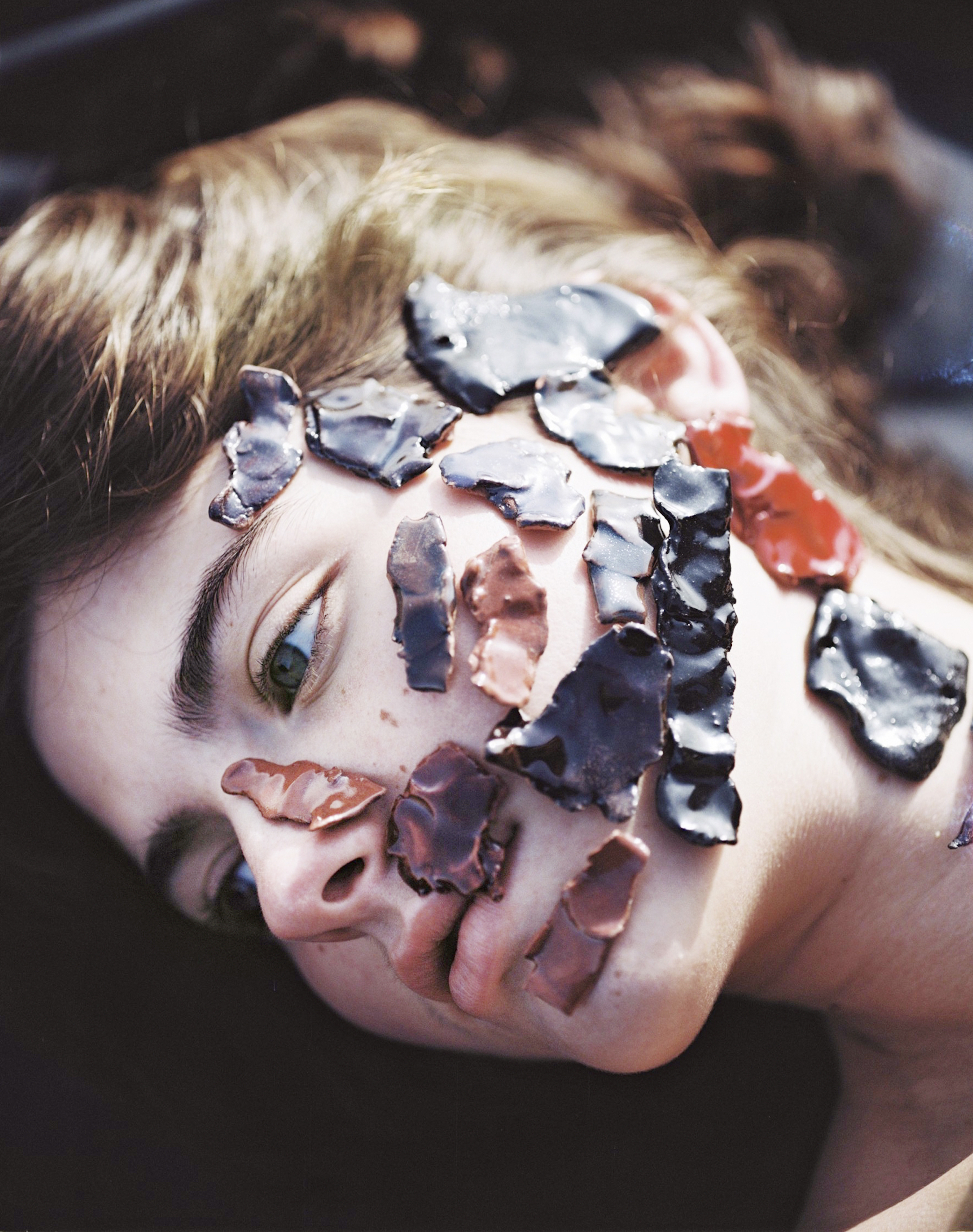 A person's face is seen with multiple pieces of painted clay attached as if makeup, the subject laying horizontally and shot with eye-level framing.