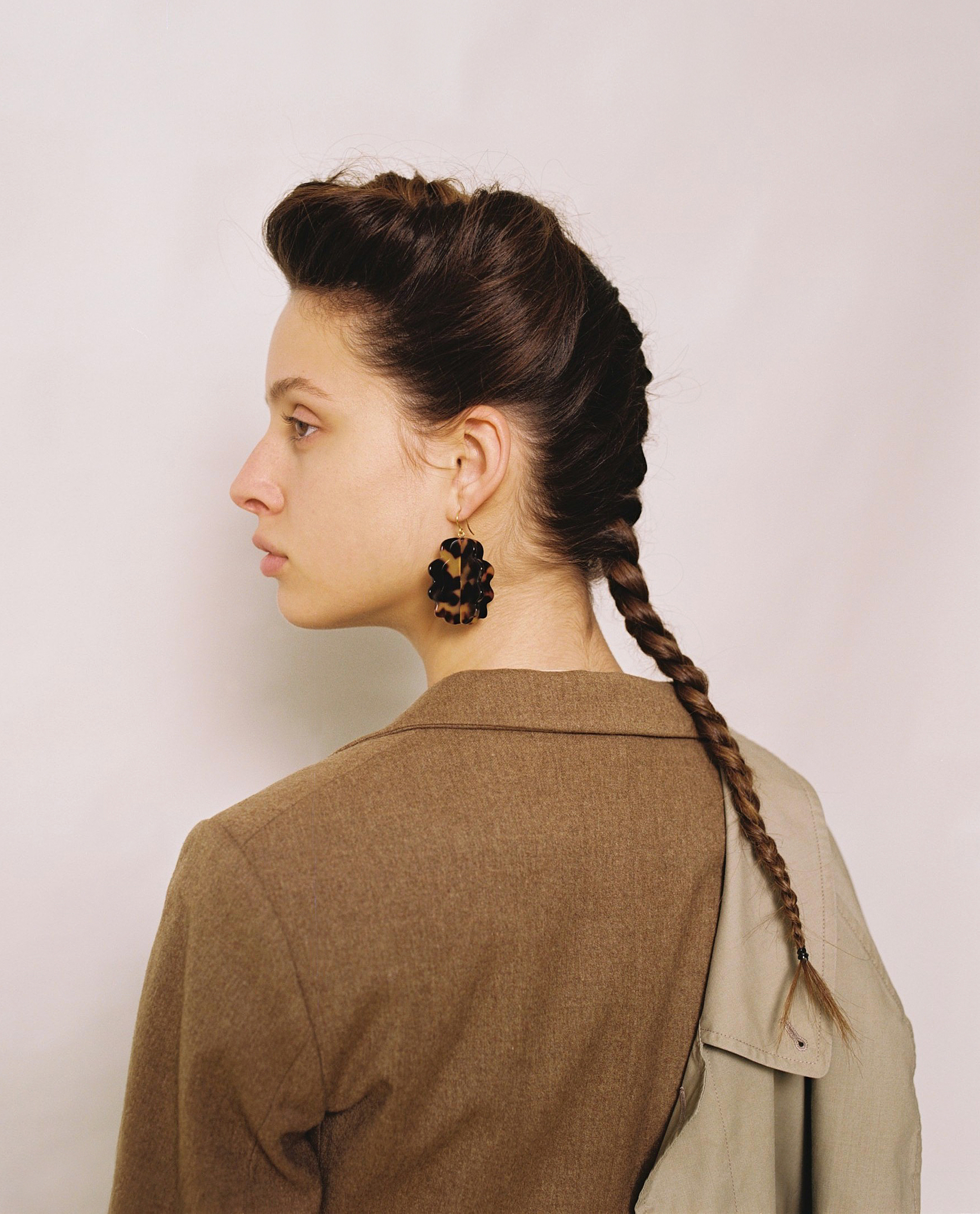 Portrait of a young woman with dark hair styled in braids wearing a brown blazer and tortoise shell earrings against a white background.
