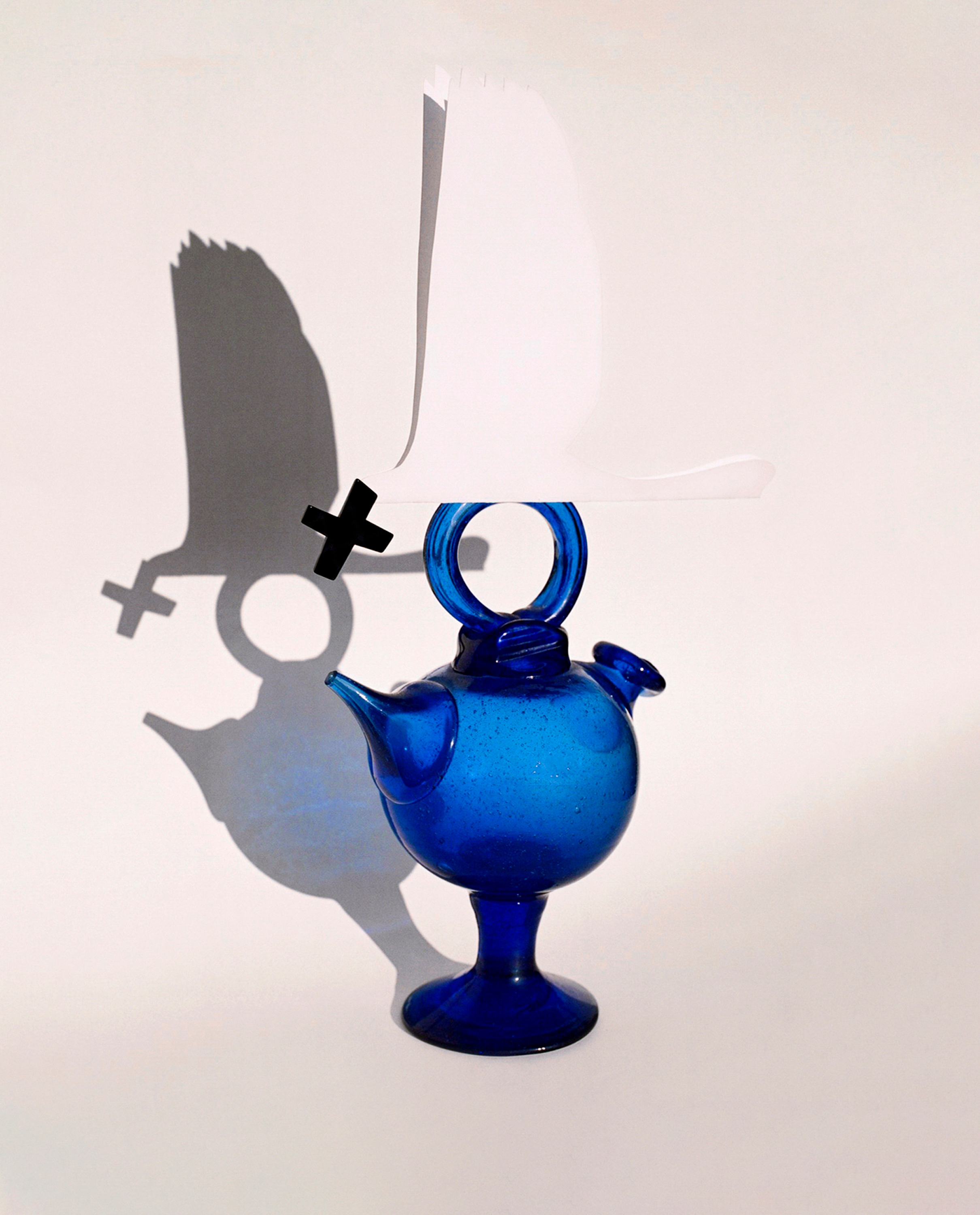 Whimsical still life featuring a blue glass teapot topped with folded white paper, casting a shadow with a Venus symbol and an X.