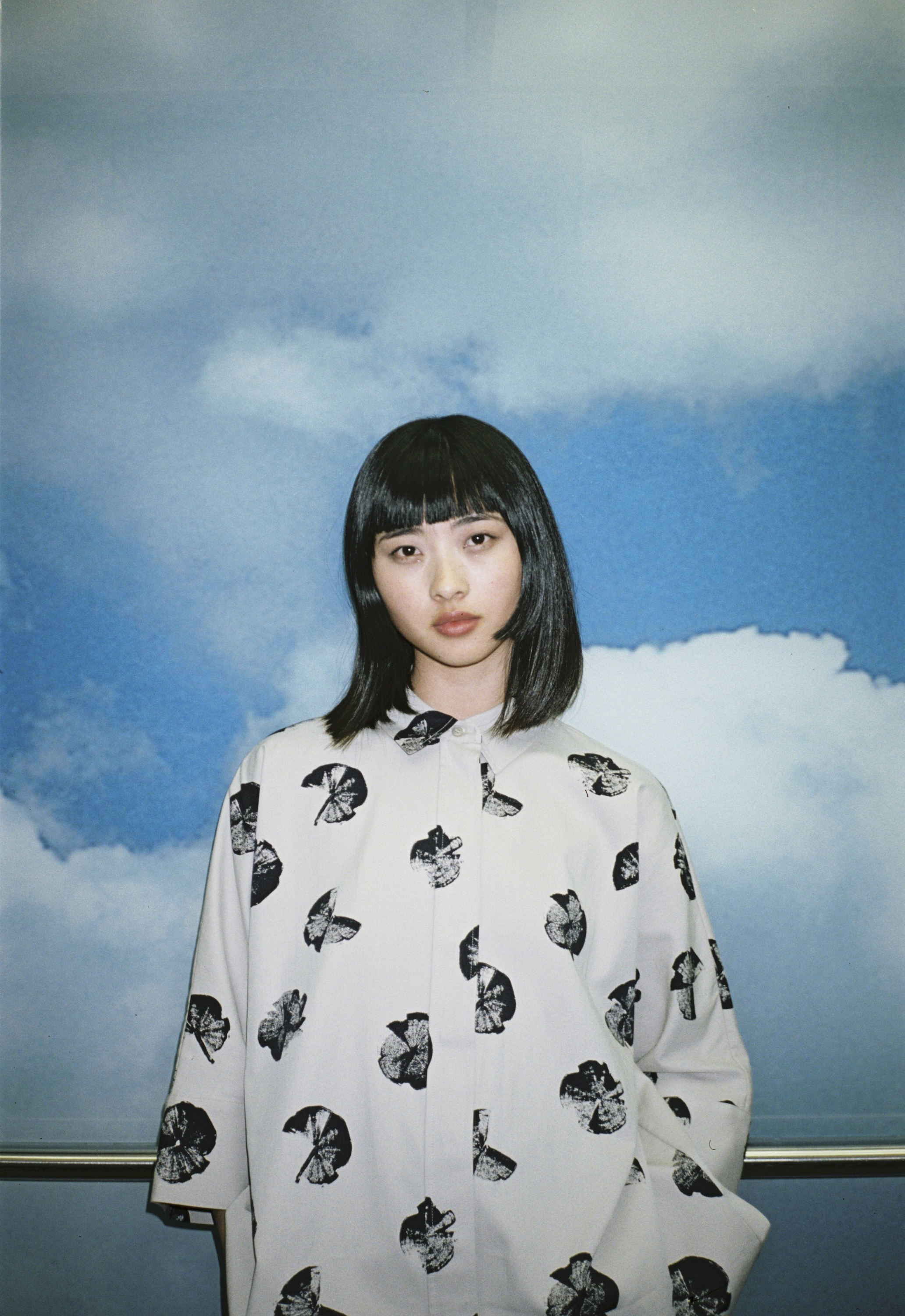 Portrait of a woman with black hair wearing a patterned shirt against a blue sky backdrop by Rita Puig-serra Costa