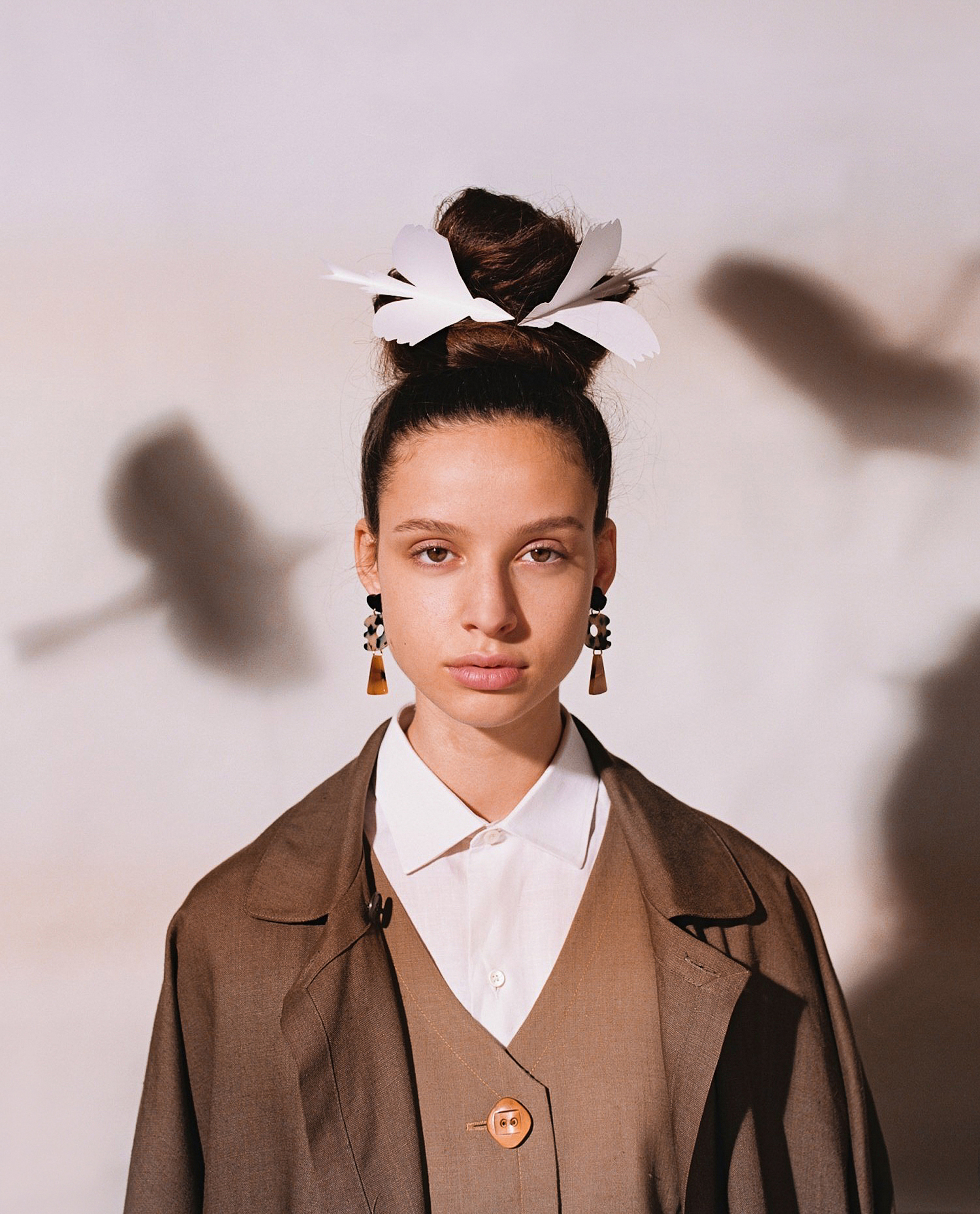 Portrait of a young woman with dark hair styled in an updo with a white flower clip, wearing a brown suit and statement earrings.