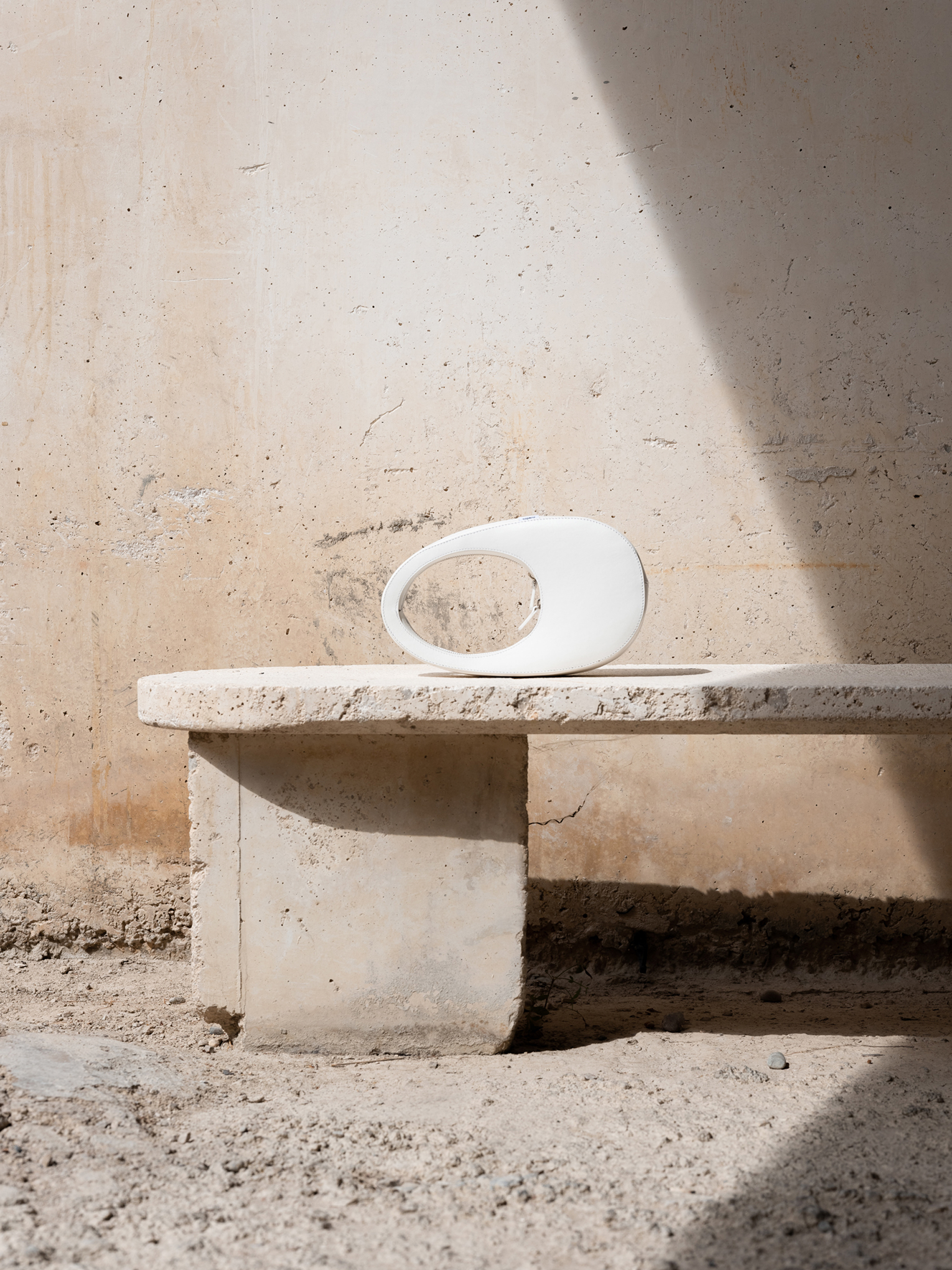 White sculptural vase sits atop a stone bench against a textured wall, illuminated by sunlight and shadows in an outdoor setting.