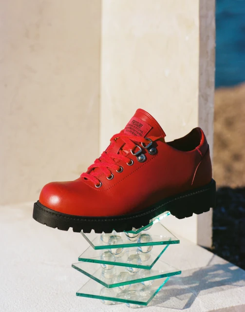 Red leather lace-up boot displayed on glass platforms against a neutral backdrop. The studio lighting highlights its smooth texture.
