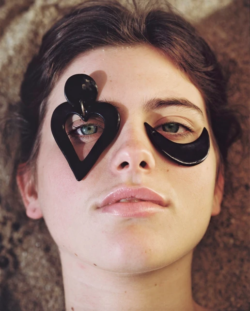 Close-up of a fair-skinned young adult woman with green eyes, wearing large black heart and crescent-shaped accessories over eyes, on sand.