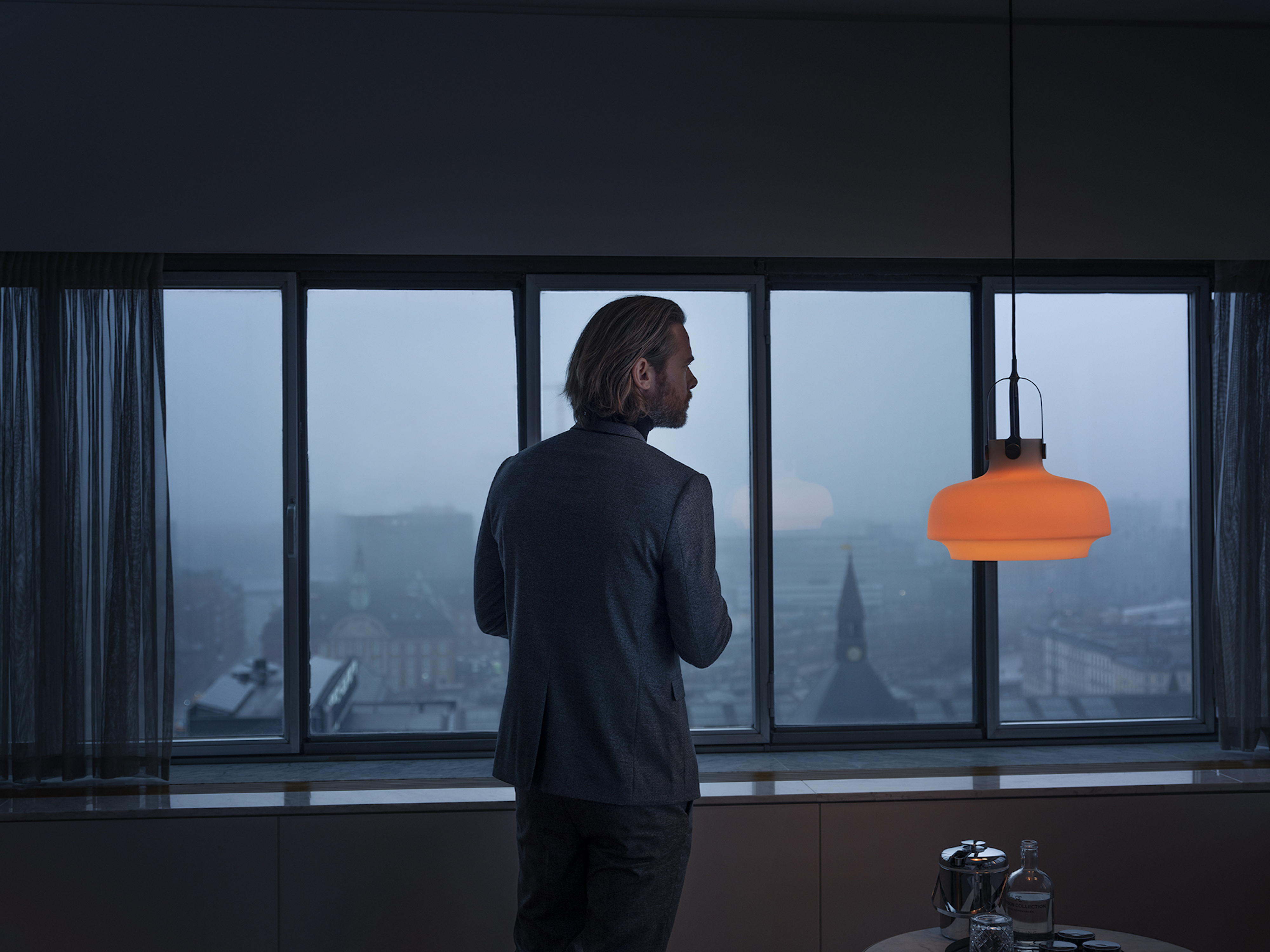Man in gray suit looks out window at misty cityscape. Indoor scene with modern lighting, table, and window view.