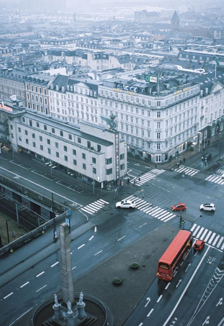 Aerial view of a city featuring buildings, streets, crosswalks, and traffic, including cars and a red double-decker bus, under an overcast sky.