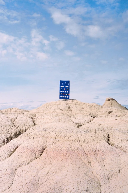 Blue and white board stands atop a white and tan desert landscape, contrasting with a bright blue sky and white clouds in the background.