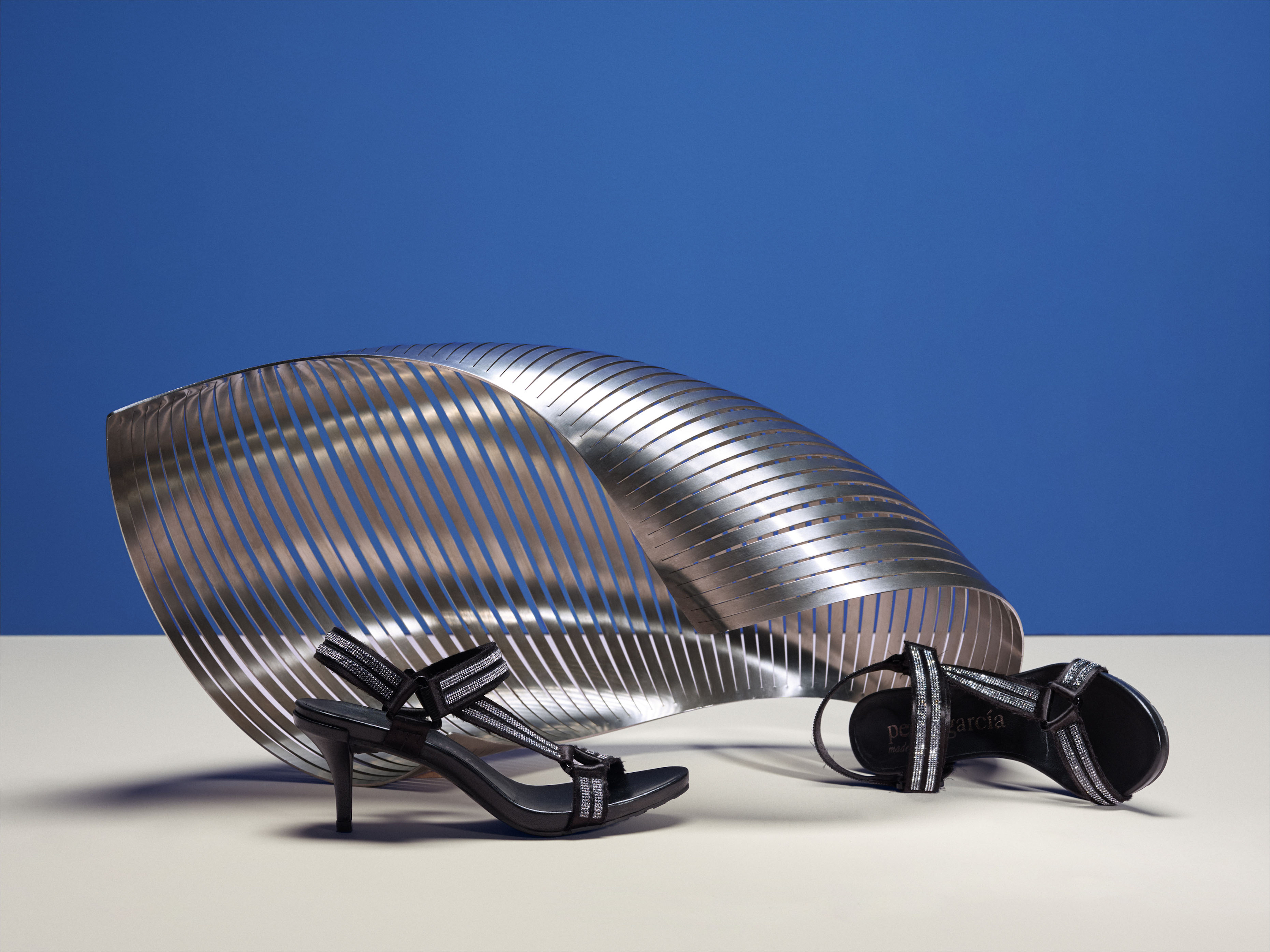 Black high-heel sandals with silver details are displayed alongside a silver sculptural element against a blue background in a studio setting.