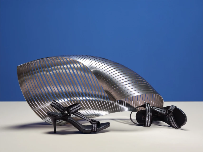 Black high-heel sandals with silver details are displayed alongside a silver sculptural element against a blue background in a studio setting.