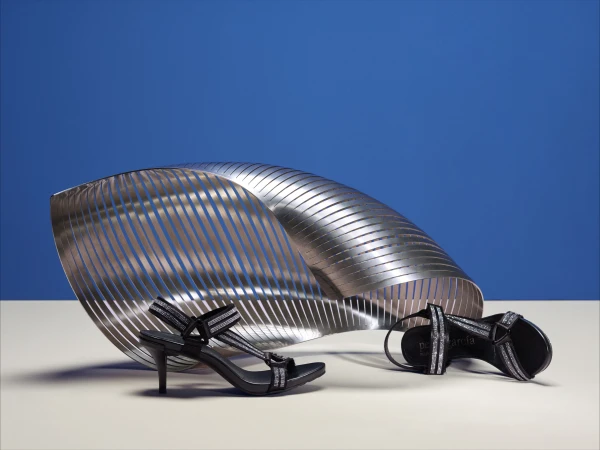 Black high-heel sandals with silver details are displayed alongside a silver sculptural element against a blue background in a studio setting.