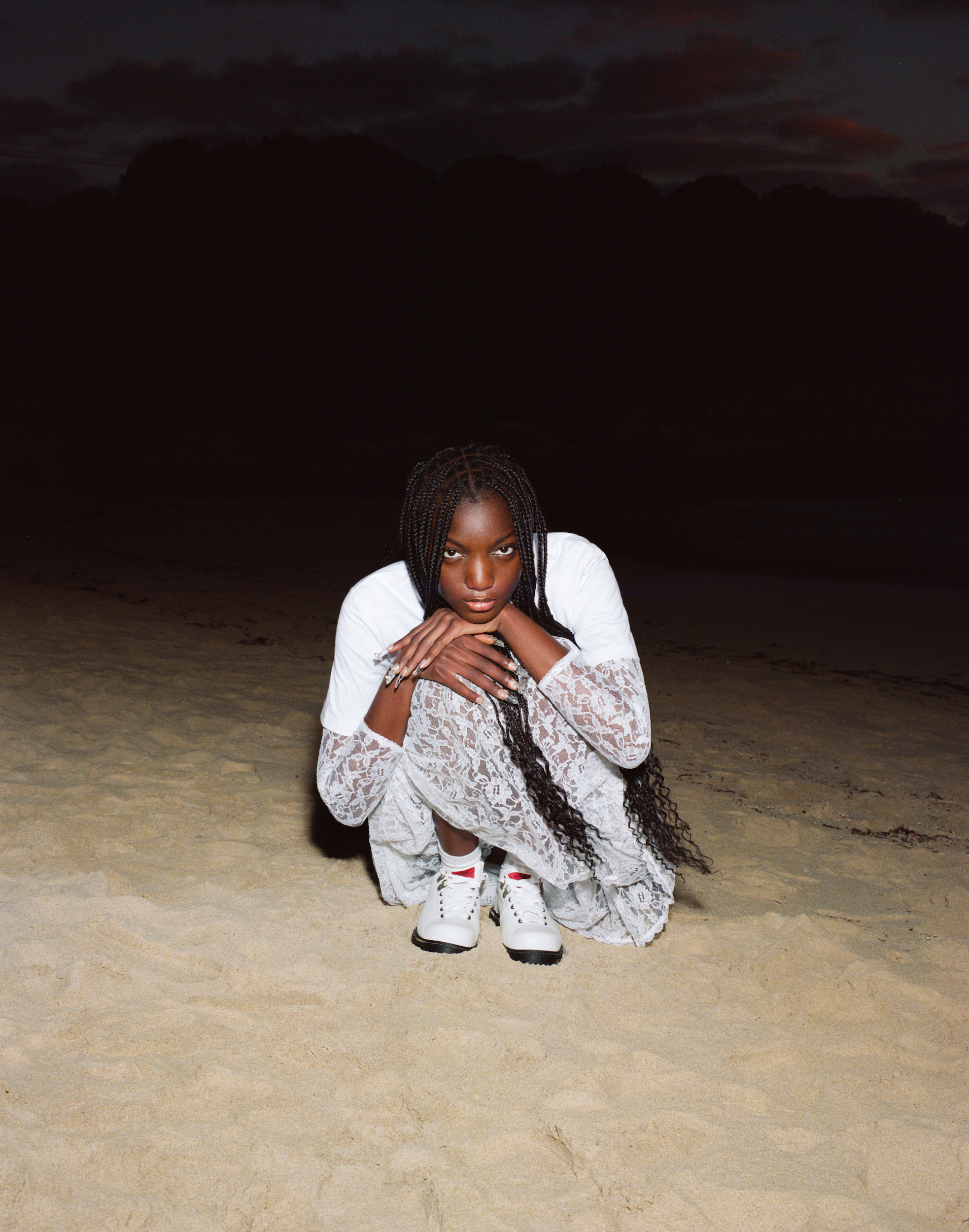 An African-descent woman sits on a sandy beach in a lace dress and sneakers with a dark sky in the background during dusk or dawn.