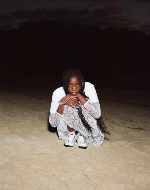 An African-descent woman sits on a sandy beach in a lace dress and sneakers with a dark sky in the background during dusk or dawn.