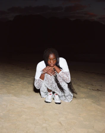 An African-descent woman sits on a sandy beach in a lace dress and sneakers with a dark sky in the background during dusk or dawn.