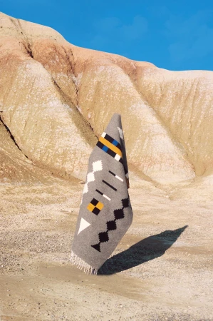 A patterned grey textile rug stands upright on a sandy desert floor against rolling arid hills and a bright blue sky.