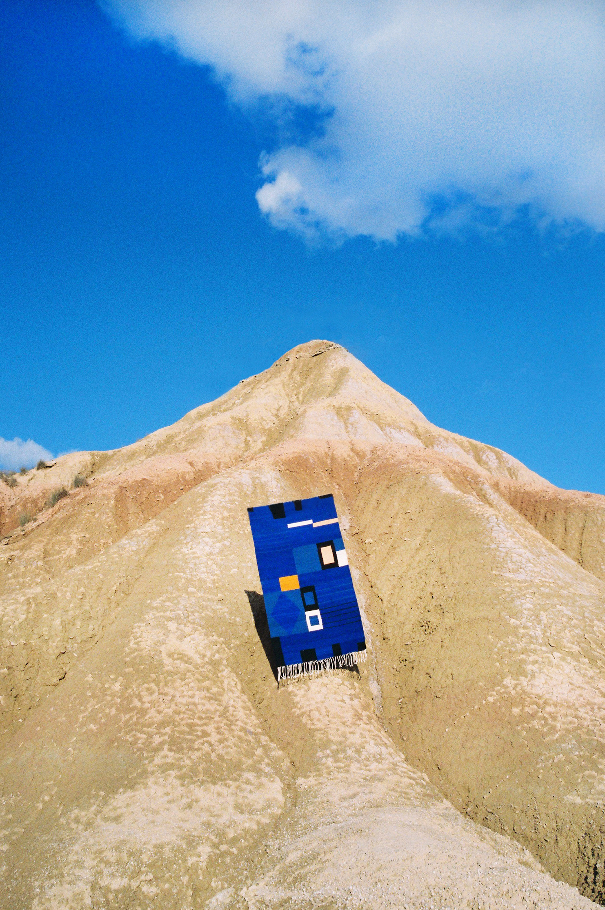 A colorful geometric rug laying on a tan-colored desert landscape against a blue sky on a bright day. Textile design on outdoor terrain.